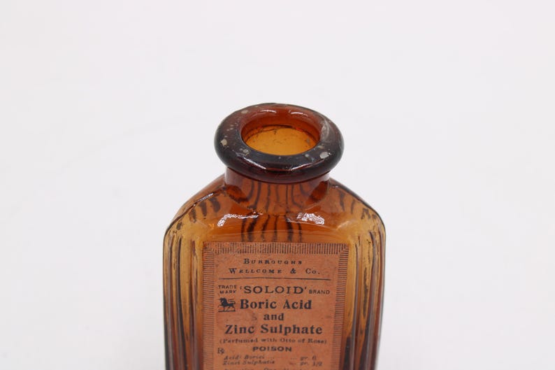 K&ouml;nnte beinhalten: Eine antike Flasche aus braunem Glas mit einem Etikett. Die Flasche hat einen dunkelbraunen Hals und einen rechteckigen K&ouml;rper mit vertikalen Rillen. Auf dem Etikett steht "Bors&auml;ure und Zinksulfat" und das Wort "GIFT".