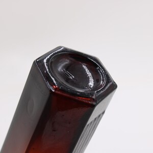 Puede incluir: Una botella de vidrio de color &aacute;mbar oscuro con una base hexagonal y un cuerpo rectangular. La botella tiene un acabado brillante y est&aacute; inclinada hacia el espectador, mostrando su forma y color sobre un fondo blanco.