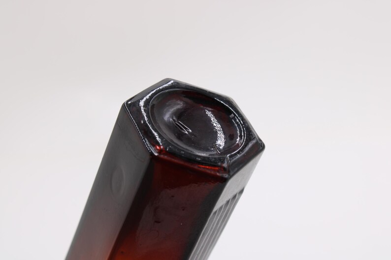 Puede incluir: Una botella de vidrio de color &aacute;mbar oscuro con una base hexagonal y un cuerpo rectangular. La botella tiene un acabado brillante y est&aacute; inclinada hacia el espectador, mostrando su forma y color sobre un fondo blanco.