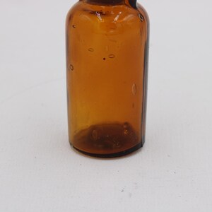 Może przedstawiać: Bursztynowa szklana butelka o zaokrąglonym korpusie i wąskiej szyjce. Butelka ma wygląd vintage, z małymi pęcherzykami powietrza widocznymi w szkle. Butelka jest pusta i stoi na białej powierzchni.