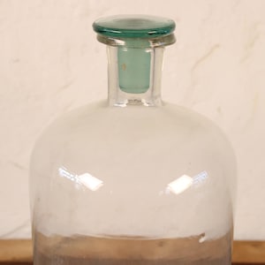 May include: A clear glass bottle with a rounded body and a narrow neck. The bottle has a green glass stopper and a brown base. The bottle is likely used for storing liquids.