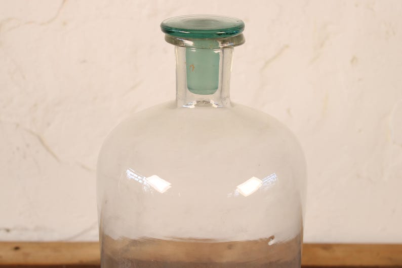 May include: A clear glass bottle with a rounded body and a narrow neck. The bottle has a green glass stopper and a brown base. The bottle is likely used for storing liquids.