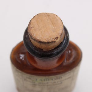 May include: Close-up of a vintage brown glass bottle with a cork stopper. The bottle has a dark brown neck and a label with faded text. The cork is light brown and shows signs of age. The bottle's shape is rounded.