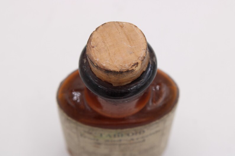 May include: Close-up of a vintage brown glass bottle with a cork stopper. The bottle has a dark brown neck and a label with faded text. The cork is light brown and shows signs of age. The bottle's shape is rounded.