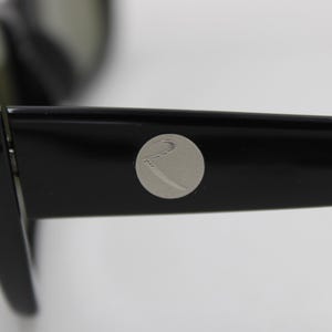 Puede incluir: Primer plano de gafas de sol negras con un emblema plateado en la patilla. Las gafas de sol tienen un dise&ntilde;o elegante y moderno con un acabado brillante. Las lentes son de color oscuro y la montura es gruesa y robusta.