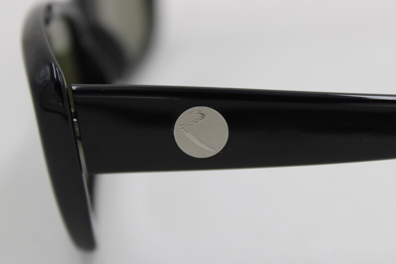 Puede incluir: Primer plano de gafas de sol negras con un emblema plateado en la patilla. Las gafas de sol tienen un dise&ntilde;o elegante y moderno con un acabado brillante. Las lentes son de color oscuro y la montura es gruesa y robusta.