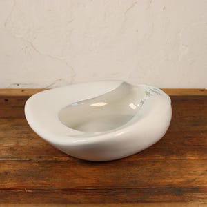 May include: A white, organically shaped ceramic bowl with a smooth, glossy finish. The bowl has a unique, asymmetrical design with a central, elongated depression. It sits on a rustic, wooden surface.