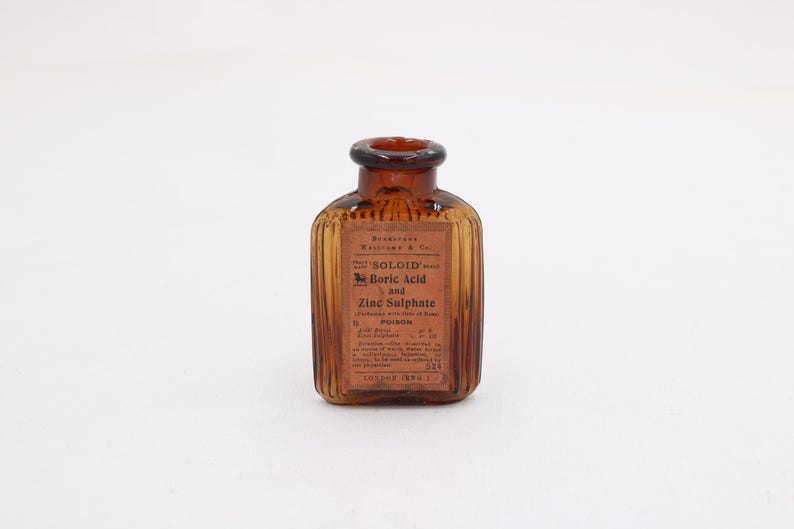 K&ouml;nnte beinhalten: Eine bernsteinfarbene Glasflasche mit braunem Etikett. Auf dem Etikett steht "SOLOID Boric Acid and Zinc Sulphate" und das Wort "POISON". Die Flasche hat ein geripptes Design und einen dunkelbraunen Verschluss.