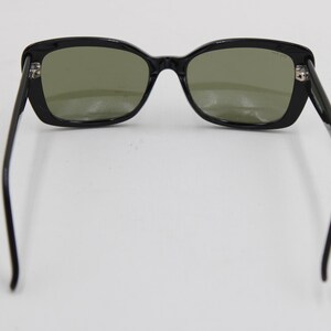 Puede incluir: Gafas de sol negras con montura rectangular y lentes tintadas en verde. Las patillas se extienden hacia afuera, creando un aspecto cl&aacute;sico y elegante. Las gafas de sol est&aacute;n sobre un fondo blanco.
