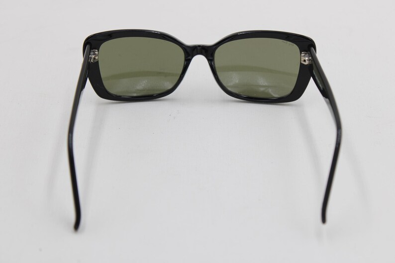 Puede incluir: Gafas de sol negras con montura rectangular y lentes tintadas en verde. Las patillas se extienden hacia afuera, creando un aspecto cl&aacute;sico y elegante. Las gafas de sol est&aacute;n sobre un fondo blanco.