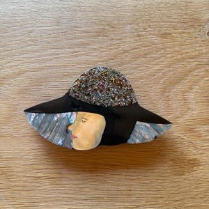 Peut inclure: Broche de style vintage représentant le profil d'une femme. Le chapeau est noir avec une couronne scintillante multicolore. Le visage est peint avec un teint clair et des lèvres rouges. La broche a un effet nacré sur le bord du chapeau.
