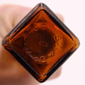 May include: Close-up of a vintage brown glass bottle base. The base is diamond-shaped with rounded corners. Embossed text and the number 6 are visible. The bottle has a dark amber color.