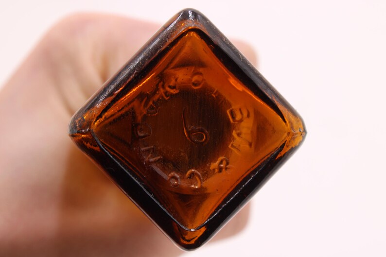 May include: Close-up of a vintage brown glass bottle base. The base is diamond-shaped with rounded corners. Embossed text and the number 6 are visible. The bottle has a dark amber color.