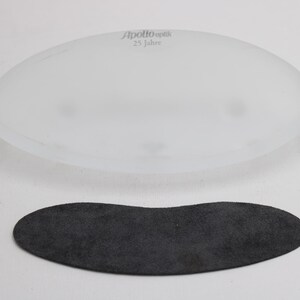 K&ouml;nnte beinhalten: Ein ovales Objekt aus mattiertem Glas mit dem Text "Apollo optik 25 Jahre" befindet sich &uuml;ber einem dunkelgrauen, Wildleder-&auml;hnlichen Oval. Die Objekte befinden sich auf einer wei&szlig;en Oberfl&auml;che. Das Glasobjekt scheint eine Linse zu sein.