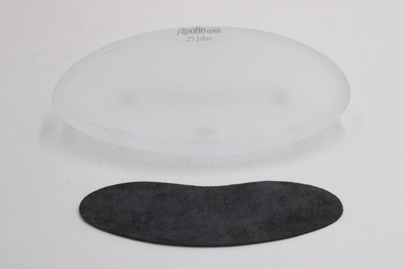 K&ouml;nnte beinhalten: Ein ovales Objekt aus mattiertem Glas mit dem Text "Apollo optik 25 Jahre" befindet sich &uuml;ber einem dunkelgrauen, Wildleder-&auml;hnlichen Oval. Die Objekte befinden sich auf einer wei&szlig;en Oberfl&auml;che. Das Glasobjekt scheint eine Linse zu sein.
