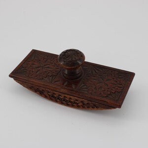 May include: A vintage wooden ink blotter with a dark brown finish. The rectangular blotter features intricate floral carvings on the top surface and a rounded base. A small, round knob is centered on the top. A classic desk accessory.