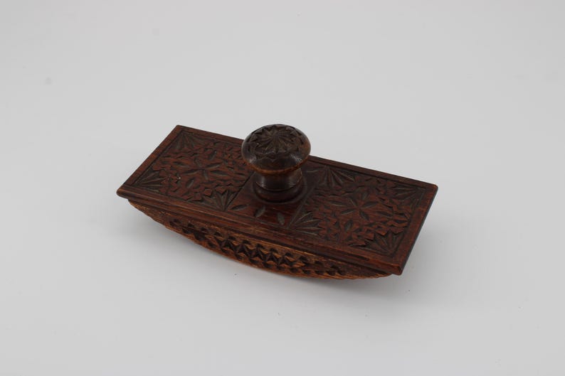 May include: A vintage wooden ink blotter with a dark brown finish. The rectangular blotter features intricate floral carvings on the top surface and a rounded base. A small, round knob is centered on the top. A classic desk accessory.