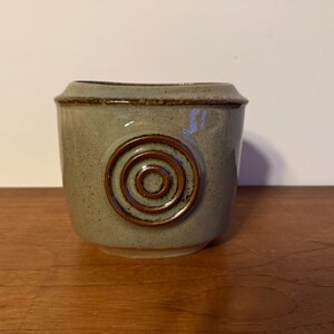 Peut inclure: Petit pot carré en céramique vert clair. Un motif circulaire en relief avec des anneaux concentriques de couleur marron foncé orne le devant. Le bord est également marron foncé. Le pot est posé sur une surface en bois.