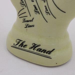 May include: A cream ceramic hand with black text and lines, featuring the words "The Hand" and "Travel Lines." The lines curve across the surface, suggesting palmistry. This decorative hand is a unique item.