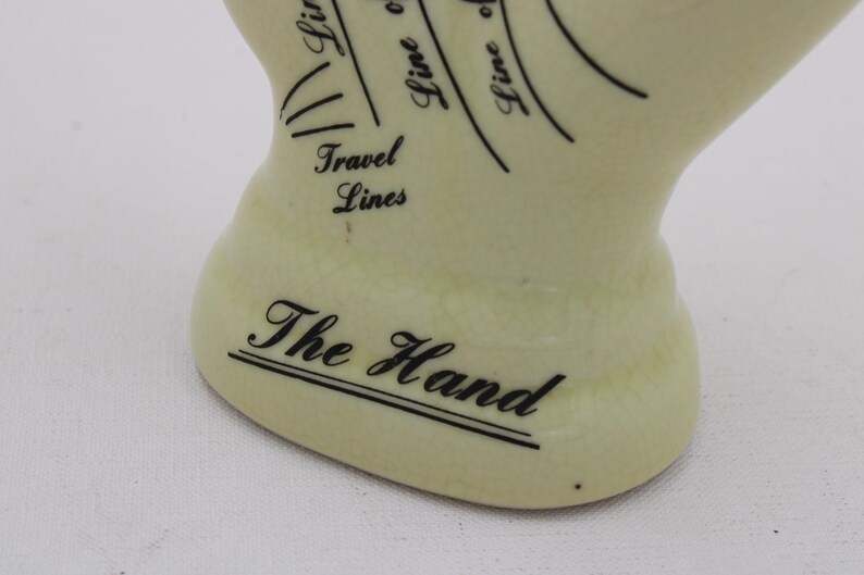 May include: A cream ceramic hand with black text and lines, featuring the words "The Hand" and "Travel Lines." The lines curve across the surface, suggesting palmistry. This decorative hand is a unique item.