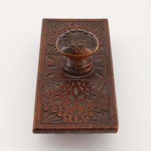 May include: A vintage wooden ink blotter with a carved floral design. The rectangular blotter has a rounded handle on top. The wood is a warm brown color, and the carved details add texture and visual interest to the piece.