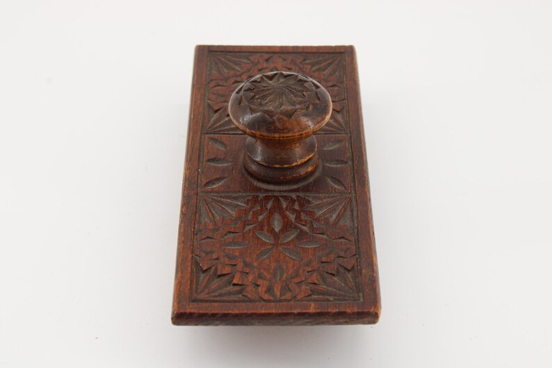 May include: A vintage wooden ink blotter with a carved floral design. The rectangular blotter has a rounded handle on top. The wood is a warm brown color, and the carved details add texture and visual interest to the piece.