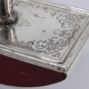 May include: Close-up of a silver ink blotter with ornate floral engravings around the edges. The blotter has a rectangular shape with a rounded, burgundy-colored base and a silver handle.
