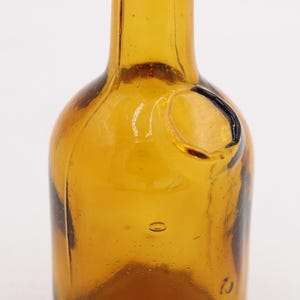 May include: Close-up of a vintage amber glass bottle. The bottle has a rounded body and a narrow neck. It features a small, circular indentation and a few air bubbles within the glass. The bottle's color is a warm, transparent amber.