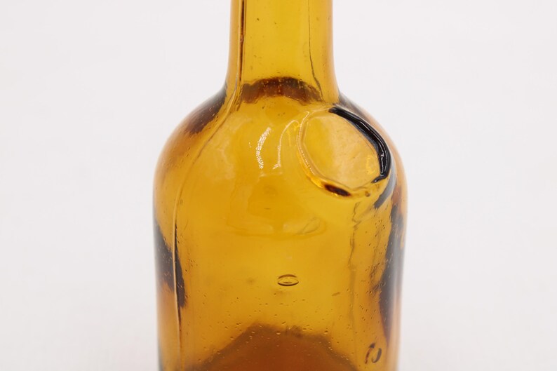 May include: Close-up of a vintage amber glass bottle. The bottle has a rounded body and a narrow neck. It features a small, circular indentation and a few air bubbles within the glass. The bottle's color is a warm, transparent amber.