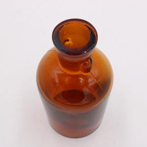 May include: Amber glass bottle with a rounded body and a narrow neck. The bottle has a small handle and a dark brown rim. The glass is transparent, allowing light to pass through. The bottle is likely used for storing liquids.
