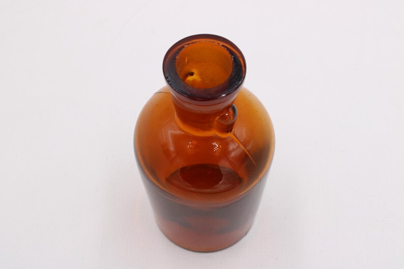 May include: Amber glass bottle with a rounded body and a narrow neck. The bottle has a small handle and a dark brown rim. The glass is transparent, allowing light to pass through. The bottle is likely used for storing liquids.