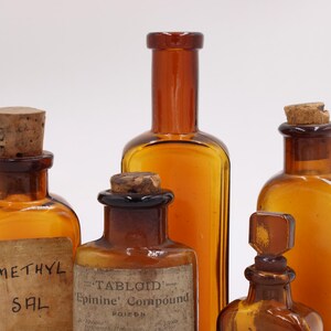 May include: An assortment of amber-colored antique apothecary bottles with cork stoppers. Some bottles have labels with faded text, including "METHYL SAL" and "TABLOID Epinine Compound POISON". The bottles vary in shape and size.