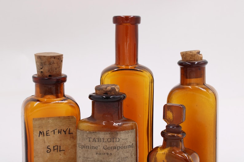 May include: An assortment of amber-colored antique apothecary bottles with cork stoppers. Some bottles have labels with faded text, including "METHYL SAL" and "TABLOID Epinine Compound POISON". The bottles vary in shape and size.