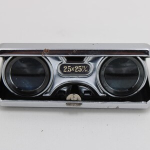 May include: A vintage pair of opera glasses with a silver-colored metal frame. The binoculars have two lenses and a central focus wheel. The text "2.5x25%" is printed on the front. The design is rectangular with a dark brown accent.