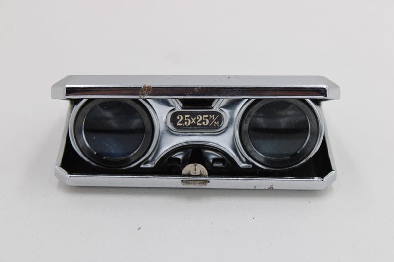 May include: A vintage pair of opera glasses with a silver-colored metal frame. The binoculars have two lenses and a central focus wheel. The text "2.5x25%" is printed on the front. The design is rectangular with a dark brown accent.