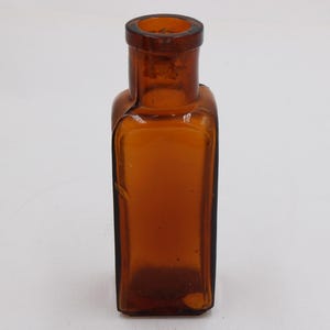 May include: An amber-colored, rectangular glass bottle with a rounded neck. The bottle has a slightly curved shape and a dark brown hue, suggesting it may be vintage or antique. The bottle is empty and sits on a white surface.
