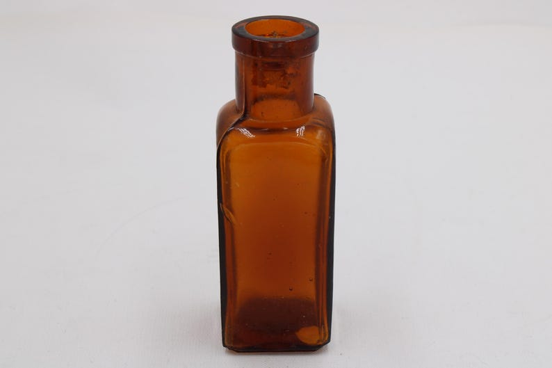 May include: An amber-colored, rectangular glass bottle with a rounded neck. The bottle has a slightly curved shape and a dark brown hue, suggesting it may be vintage or antique. The bottle is empty and sits on a white surface.