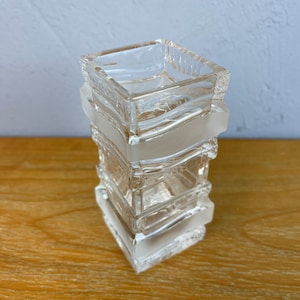 May include: A clear glass vase with a square shape and a textured design. The vase is made of multiple stacked layers of glass.