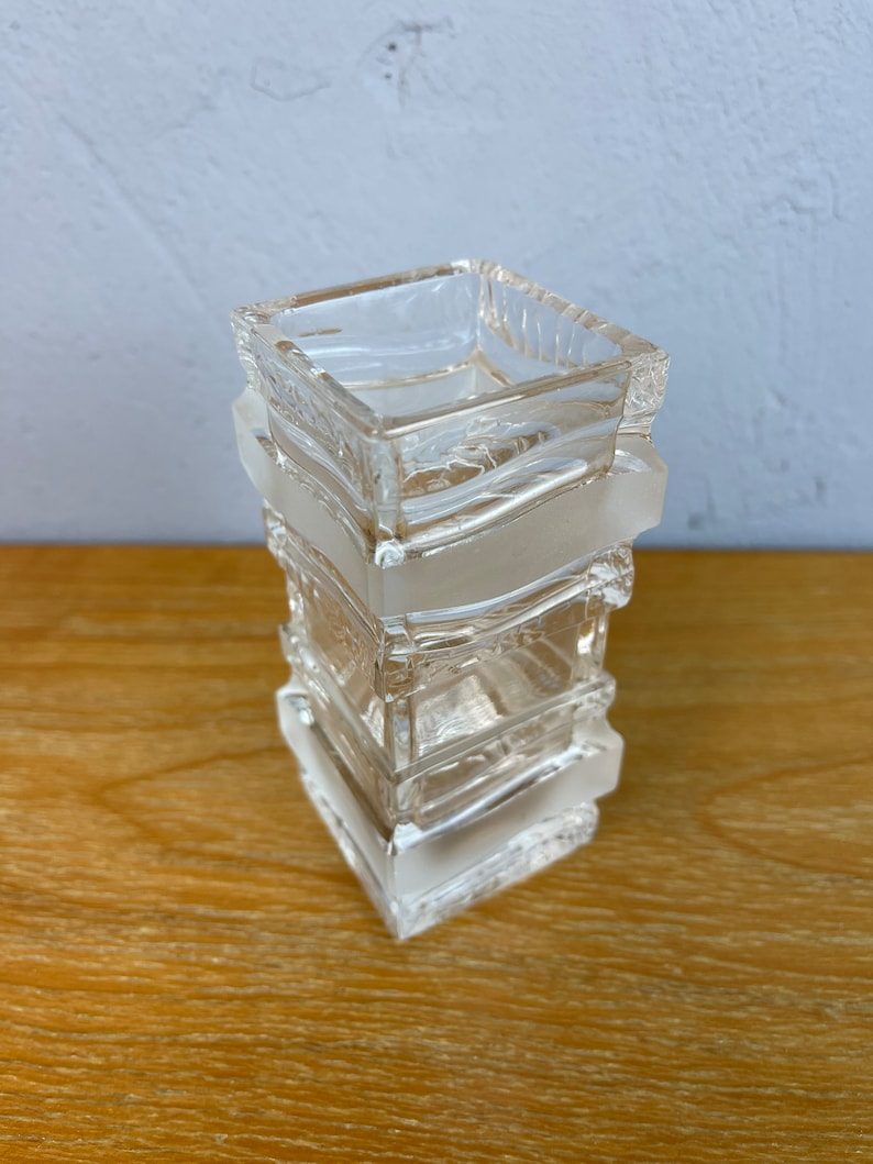 May include: A clear glass vase with a square shape and a textured design. The vase is made of multiple stacked layers of glass.