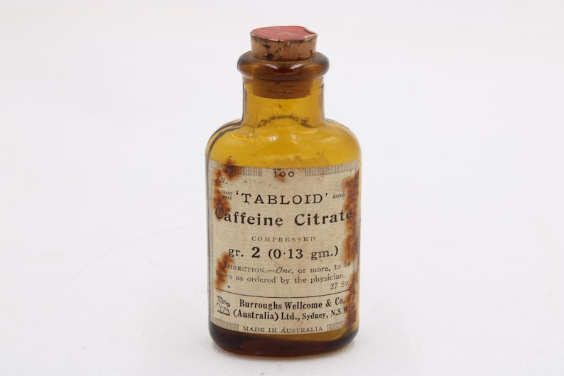May include: An old amber glass bottle with a cork stopper. The bottle has a label with the text "TABLOID Caffeine Citrate" and "gr. 2 (0.13 gm.)". The label also includes the manufacturer's name and "MADE IN AUSTRALIA".