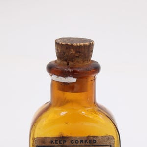 May include: An amber glass bottle with a cork stopper. The bottle has a rounded shape and a label that reads "KEEP CORKED". The cork is light brown, and the bottle has a vintage appearance.