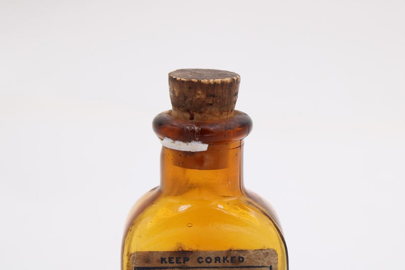 May include: An amber glass bottle with a cork stopper. The bottle has a rounded shape and a label that reads "KEEP CORKED". The cork is light brown, and the bottle has a vintage appearance.