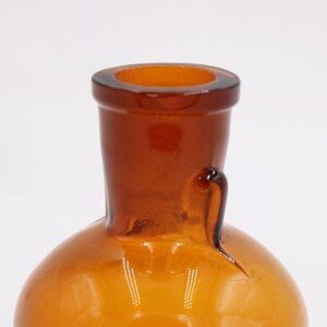 May include: Close-up of a vintage amber glass bottle. The bottle has a rounded base and a long, tapered neck with a flared rim. A small handle is attached to the side of the neck. The glass has a warm, golden-brown color.