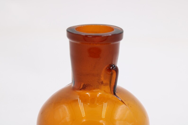 May include: Close-up of a vintage amber glass bottle. The bottle has a rounded base and a long, tapered neck with a flared rim. A small handle is attached to the side of the neck. The glass has a warm, golden-brown color.