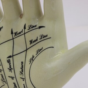 May include: A cream-coloured ceramic hand with black lines and text indicating palmistry features. The text includes "Marriage Lines," "Heart Line," "Head Line," and "Life Line." The hand is a decorative item.