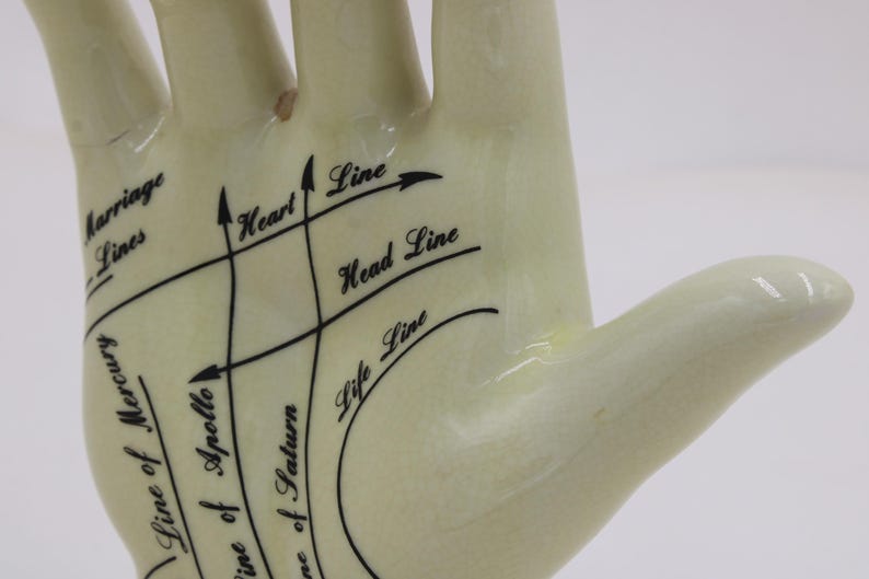 May include: A cream-coloured ceramic hand with black lines and text indicating palmistry features. The text includes "Marriage Lines," "Heart Line," "Head Line," and "Life Line." The hand is a decorative item.