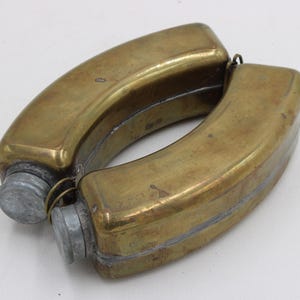 May include: A vintage brass flask with a curved, double-sided design. The flask has a tarnished, aged appearance, with two metal screw-on caps. The overall shape is reminiscent of a figure eight, suggesting it may have been designed to be carried on a belt.