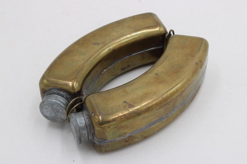 May include: A vintage brass flask with a curved, double-sided design. The flask has a tarnished, aged appearance, with two metal screw-on caps. The overall shape is reminiscent of a figure eight, suggesting it may have been designed to be carried on a belt.