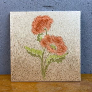 Puede incluir: Un azulejo de cerámica vintage con un diseño floral. El azulejo presenta dos flores de amapola naranja con hojas verdes sobre un fondo blanco con un glaseado marrón moteado.