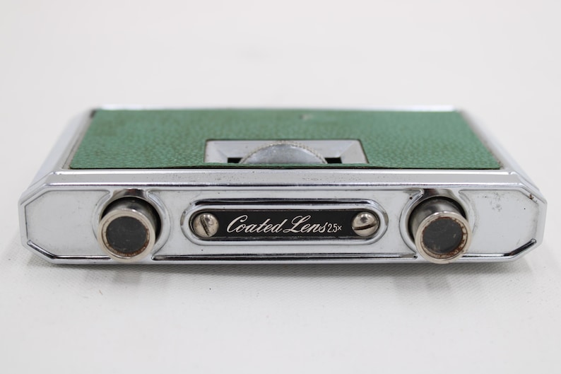 May include: A vintage pair of silver-toned opera glasses with a green textured top. The binoculars have a central focus wheel and the text "Coated Lens 2.5x" is visible on the front.